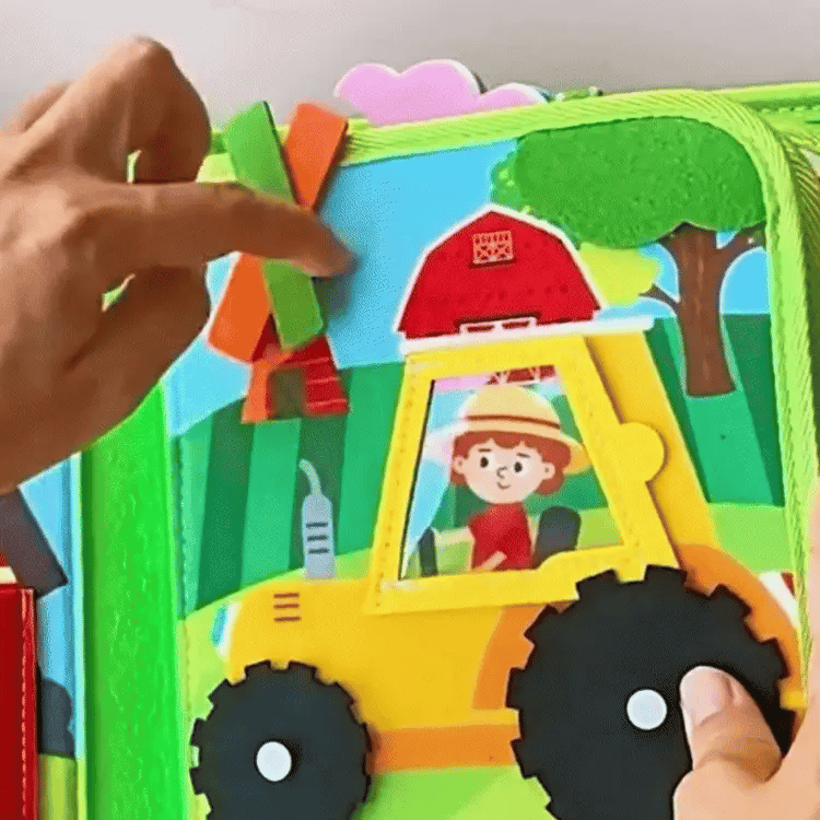 Detaljan prikaz interaktivne stranice senzorne knjige od filca koja prikazuje žuti traktor s pomičnim crnim kotačima i vozačem. Ruka pokazuje na vjetrenjaču s narančastim i zelenim krakovima, dok se u pozadini nalaze motivi farme poput crvene štale i drveća. Slika odgovara datoteci pod nazivom "a51-ezgif.com-video-to-gif-converter_a1a083d2-1749-4323-bec5-d0d1e1a5ae82".