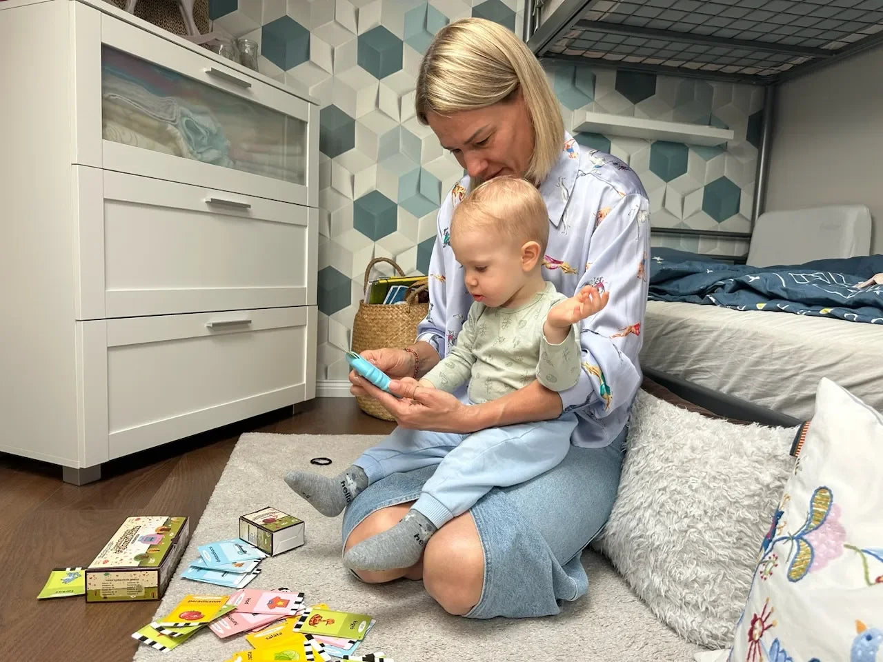 Majka i dijete u interaktivnoj igri s plavim KinderSpeech čitačem i edukativnim karticama na podu sobe, potičući razvoj govora i rano učenje.